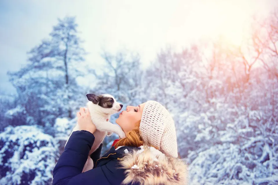 Une jeune femme en bonnet de laine embrasse un chiot noir et blanc qu'elle tient à bout de bras dans un paysage enneigé